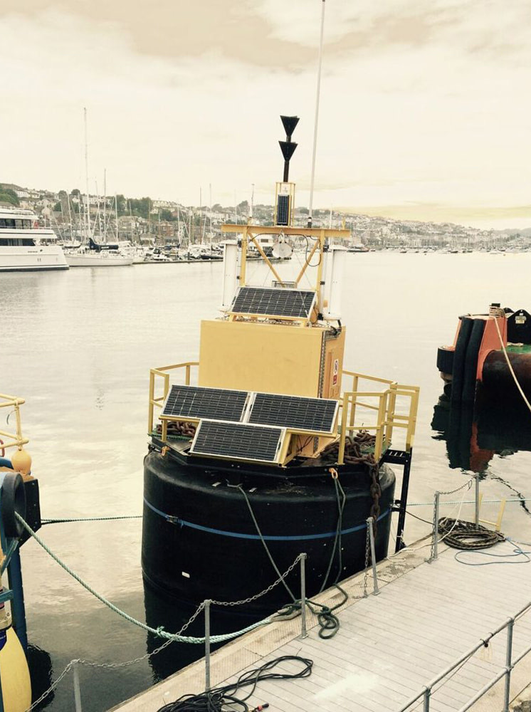 A marine buoy with a GRP housing and solar panels, moored in a harbour
